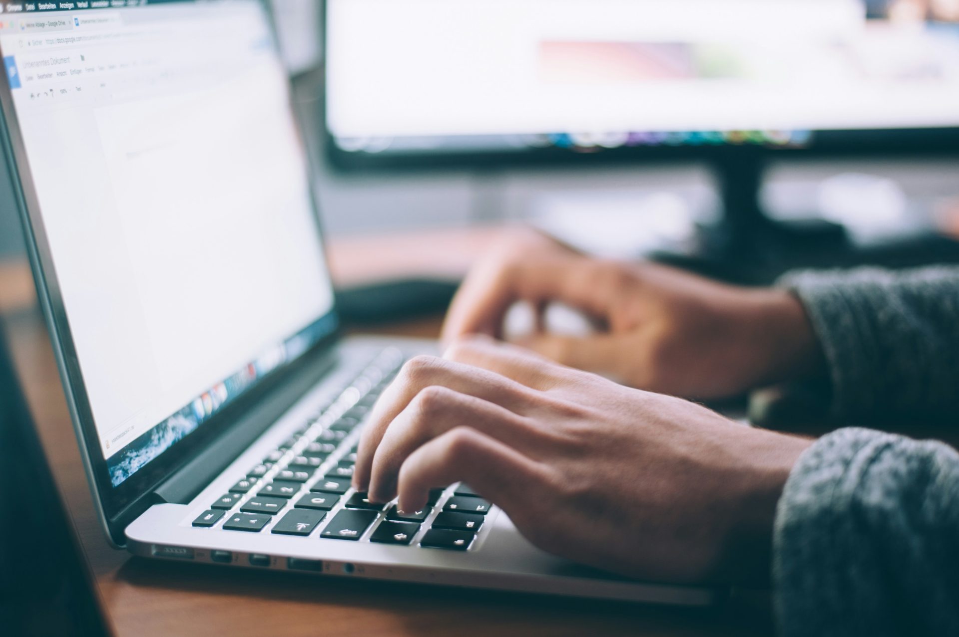 Hands typing on laptop near large monitor.